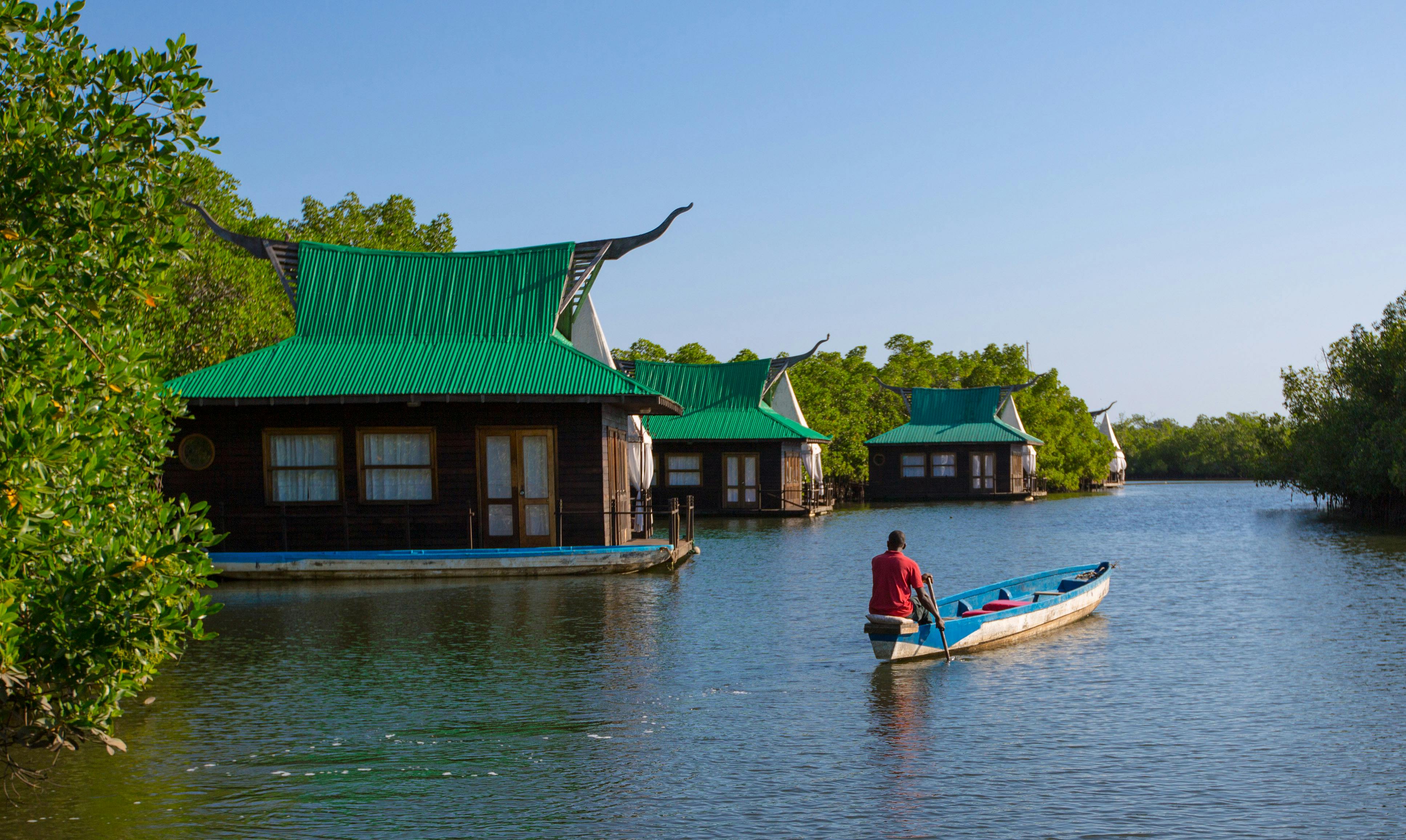 Green-roofed wooden houseboats on calm water with a man in a canoe among mangroves