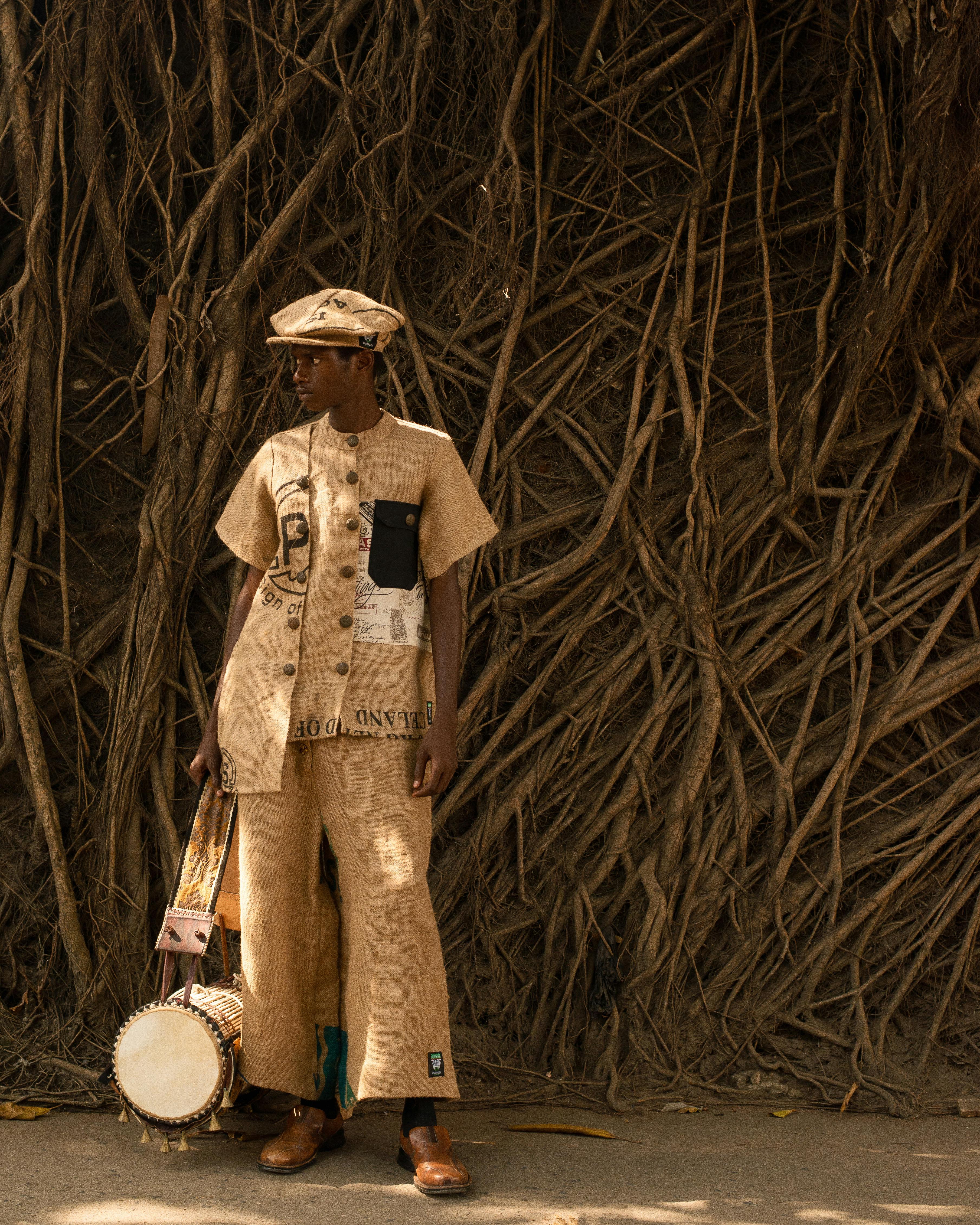 Young man in a patterned burlap-style outfit and flat cap standing beside a drum against tree roots
